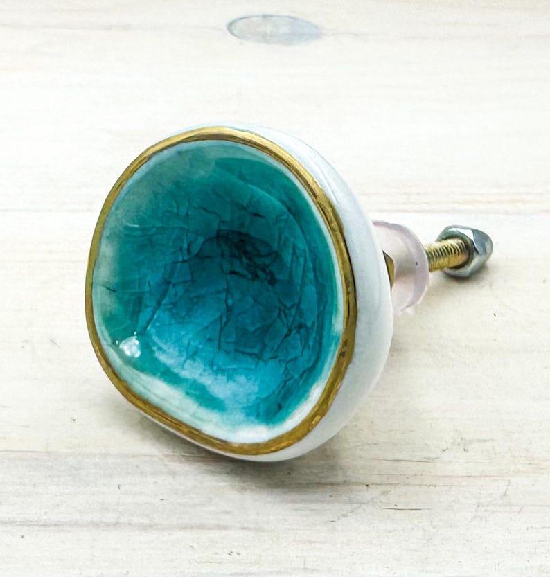 May include: A round cabinet knob with a concave center featuring a turquoise crackle glaze finish. The outer edge is painted with a metallic gold border, while the exterior side is white. A metal bolt and nut extend from the back of the knob.