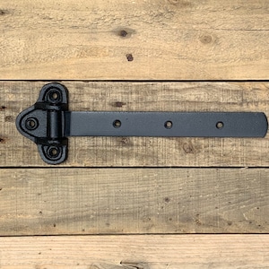 May include: Black metal hinge with four holes and a rounded edge. The hinge is attached to a wooden surface.