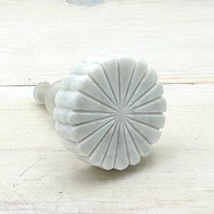 May include: A white marble bottle stopper with a carved, fan-like design. The stopper has a rounded, ribbed body and a cylindrical stem. The product is made of stone and is designed to fit into a bottle.