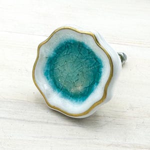 May include: A ceramic cabinet knob with a white base, teal centre, and gold trim. The knob has an organic shape and a metal screw. The teal centre has a crackle effect. The knob is suitable for cabinets and drawers.