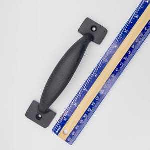 May include: Black metal door handle with a rounded shape and square ends. The handle is 12.7 cm long and is measured against a ruler.