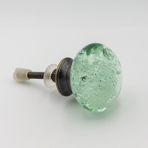 May include: A green glass cabinet knob with a brass base and a screw. The glass is filled with small air bubbles.