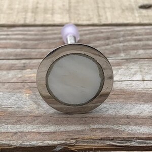 K&ouml;nnte beinhalten: Ein silberfarbener Flaschenverschluss mit einer wei&szlig;en, runden, dekorativen Oberseite. Der Verschluss hat eine glatte, gl&auml;nzende Oberfl&auml;che.