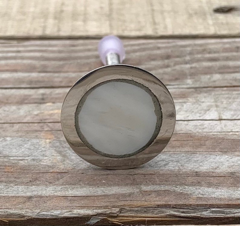 K&ouml;nnte beinhalten: Ein silberfarbener Flaschenverschluss mit einer wei&szlig;en, runden, dekorativen Oberseite. Der Verschluss hat eine glatte, gl&auml;nzende Oberfl&auml;che.