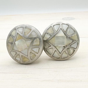 Puede incluir: Dos tiradores de armario redondos, de color plateado. Cada tirador presenta una forma de diamante central y formas triangulares circundantes con incrustaciones de nácar iridiscente. Los tiradores están sobre un fondo de madera clara.