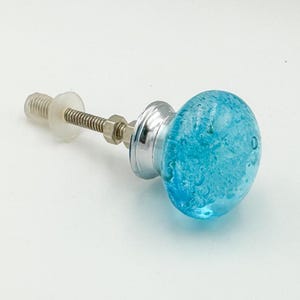 May include: A round cabinet knob featuring a bright blue glass head with internal bubble textures. The knob is attached to a silver-toned metal base and a threaded metal bolt with a small nut and a translucent plastic washer for installation.
