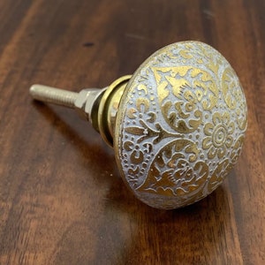 May include: A white ceramic knob with a gold floral design. The knob has a silver screw.
