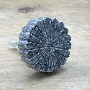 May include: A grey stone knob with a circular, sunburst-like design. The knob has a smooth, polished finish and a small, clear stem.