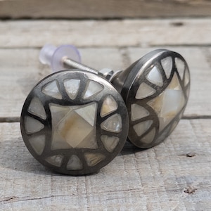 May include: Two round, silver-toned cabinet knobs with a geometric design. The knobs are made of mother-of-pearl and have a black metal frame.