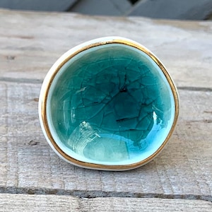 May include: A round ceramic cabinet knob with a teal crackle glaze center and a gold rim. The knob has a white base and is designed for furniture. The knob is a decorative accent for drawers, cabinets, or other home decor projects.