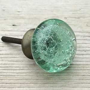 May include: A light green glass cabinet knob with a silver metal base and a screw. The glass has small air bubbles throughout.