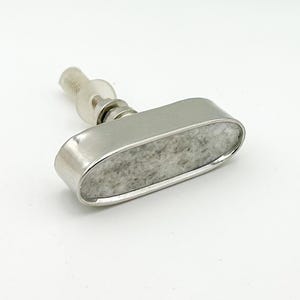 May include: An oval-shaped cabinet knob featuring a polished silver-tone metal frame surrounding a textured, light gray stone inlay. The hardware includes a threaded metal bolt, a clear plastic washer, and a metal nut for secure installation.