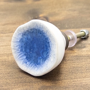 May include: White ceramic cabinet knob with a blue, crackled glaze in the centre. The knob has a silver screw and a clear plastic washer.