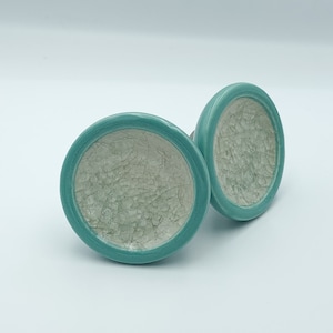 May include: Two ceramic cabinet knobs. Each knob has a round shape with a turquoise rim and a white, crackled interior. The knobs are designed for furniture and home decor.
