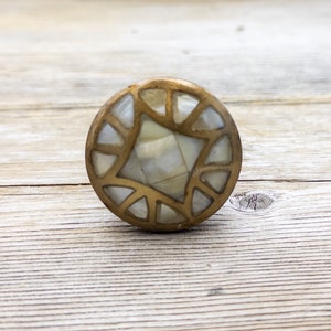 May include: A circular cabinet knob in a brass finish, inlaid with mother-of-pearl in a star and segmented design. The knob is set against a light wooden surface. The knob's design is geometric and decorative.