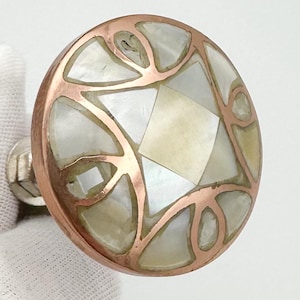 May include: A round cabinet knob with a copper-coloured metal rim and a mother-of-pearl inlay. The inlay features a geometric design with a central diamond shape and surrounding irregular shapes. The knob has a silver-coloured stem.