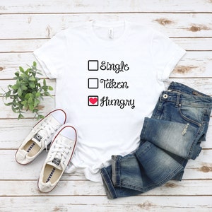 May include: White t-shirt with the text "Single, Taken, Hungry" in black. The word "Hungry" has a red heart symbol. The shirt is paired with blue jeans and white sneakers.