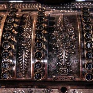 May include: Close-up of an antique cash register with ornate metalwork. The register features round price buttons with dollar amounts, from $1 to $80. The design includes decorative floral and geometric patterns.