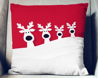 May include: Red and white throw pillow with a design of four reindeer peeking over a snowy hill.