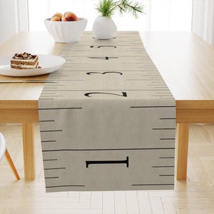 May include: A beige table runner with a ruler design. Black numbers and markings indicate measurements. The runner is placed on a wooden table. A white bowl with fruit, a vase with greenery, and a plate with cake are also on the table.