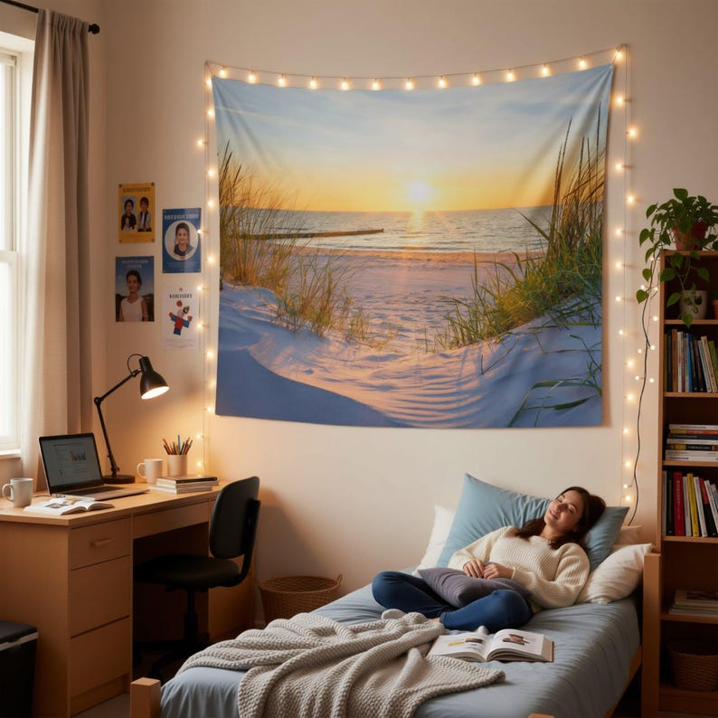 A woman is lying on a bed in a cozy bedroom, with a large tapestry of a beach scene hanging on the wall behind her. The tapestry features a sunset over the ocean and grassy dunes. The room also includes a desk with a laptop, a potted plant, and various books and decor items.