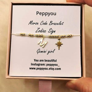 May include: A gold-coloured Morse code bracelet with a Gemini girl zodiac sign charm. The bracelet is presented in a gift box with the text "Peppyou" and "You are beautiful". The bracelet is strung on a thin, light-coloured cord.