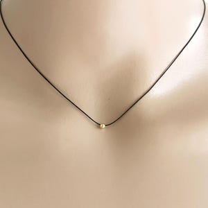 May include: A delicate necklace featuring a thin black cord and a single small, round, polished gold bead centred on the strand. The minimalist design rests against the neck, highlighting the contrast between the dark cord and the gold accent.