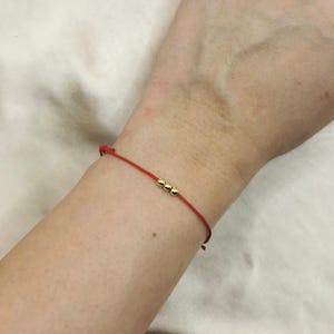 May include: A thin red string bracelet featuring three small, polished gold-toned spherical beads centered on the cord. The bracelet is designed with a simple, minimalist construction, showcasing the vibrant red cord against the metallic gold beads.
