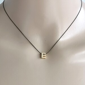 May include: A gold-toned letter E charm pendant suspended on a thin black cord necklace. The letter E is block-shaped with a metallic finish, centred on the cord. The necklace rests against the neck, showcasing the simple design of the pendant.