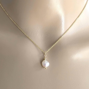 May include: A delicate gold chain necklace featuring a single, small, irregular white pearl pendant. The pearl is suspended from a simple gold jump ring, creating a minimalist look that rests against the collarbone area on a neutral base.
