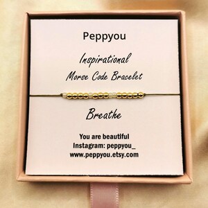 May include: A pink box displaying a white card with the text "Peppyyou Inspirational Morse Code Bracelet Breathe". A gold and white beaded bracelet is visible. The card also includes the text "You are beautiful".