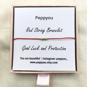 May include: A red string bracelet with three small gold-toned beads centred on a white display card inside a light pink box. The card features the text Peppyou, Red String Bracelet, Good Luck and Protection, You are beautiful, Instagram: peppyou_, and www.peppyou.etsy.com.