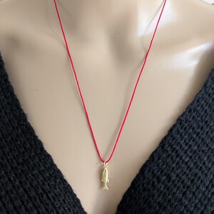 May include: A gold-coloured fish pendant suspended from a thin, vibrant red cord necklace. The small, detailed fish charm hangs vertically from a simple gold jump ring, contrasting against the bright red string and the textured black fabric.