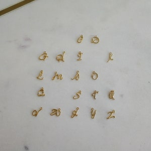 K&ouml;nnte beinhalten: Eine Sammlung kleiner, goldfarbener Buchstabenanh&auml;nger in Schreibschrift, jeweils mit einer Schlaufe zum Befestigen an Schmuck. Die Buchstaben sind auf einer wei&szlig;en Marmoroberfl&auml;che angeordnet, die die filigranen Details zeigt.