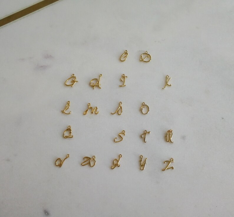 K&ouml;nnte beinhalten: Eine Sammlung kleiner, goldfarbener Buchstabenanh&auml;nger in Schreibschrift, jeweils mit einer Schlaufe zum Befestigen an Schmuck. Die Buchstaben sind auf einer wei&szlig;en Marmoroberfl&auml;che angeordnet, die die filigranen Details zeigt.