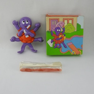 May include: A purple octopus figurine wearing an orange shirt, holding a white object, and a matching box with an illustration of an octopus at a pool. Also included are two toothbrushes in a clear plastic wrapper.