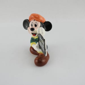 Peut inclure: Figurine en c&eacute;ramique de Mickey Mouse portant un b&eacute;ret orange, une chemise verte et jaune et des chaussures marron. La figurine tient un panneau noir et blanc. La figurine a des oreilles noires et une bouche rouge.