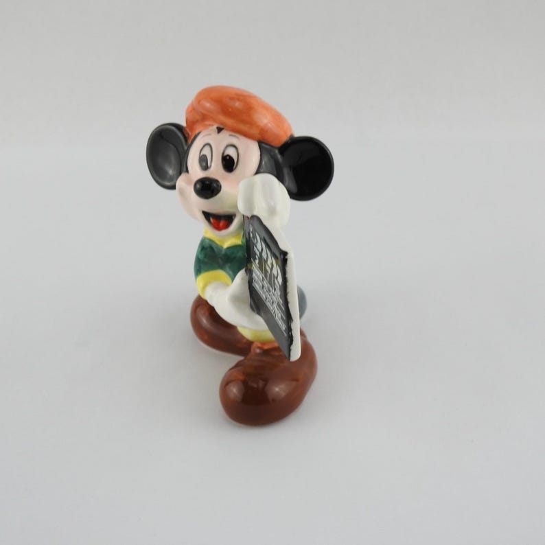 Peut inclure: Figurine en c&eacute;ramique de Mickey Mouse portant un b&eacute;ret orange, une chemise verte et jaune et des chaussures marron. La figurine tient un panneau noir et blanc. La figurine a des oreilles noires et une bouche rouge.