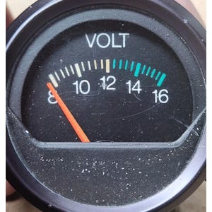 May include: A circular analog voltmeter gauge with a black face and white text reading VOLT. The scale ranges from 8 to 16, featuring white tick marks for 8 to 12 and green tick marks for 12 to 16. A bright orange needle points to the 8 mark.
