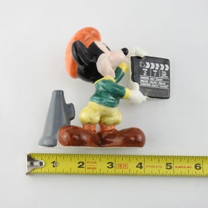 Peut inclure: Figurine de Mickey Mouse tenant un clap de cin&eacute;ma. La figurine porte une veste verte, un pantalon jaune et des chaussures marron. Un m&eacute;gaphone gris est &agrave; gauche. Le clap de cin&eacute;ma porte l'inscription "Mickey Mouse".