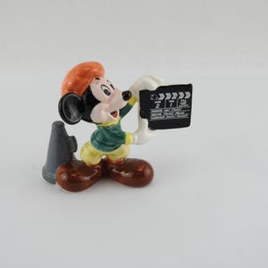 Peut inclure: Figurine de Mickey Mouse tenant un clap de cin&eacute;ma. La figurine porte une veste verte, un pantalon jaune et des chaussures marron. Elle a un chapeau rouge et un m&eacute;gaphone gris. Le clap de cin&eacute;ma contient du texte.