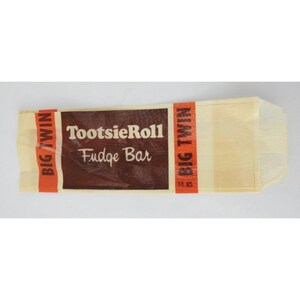 May include: A TootsieRoll Fudge Bar wrapper. The wrapper is a light tan color with a brown rectangular section in the middle. The text "TootsieRoll Fudge Bar" is printed in white. The words "Big Twin" are printed in red on the ends.
