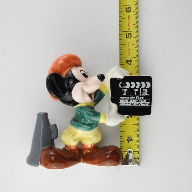 Peut inclure: Figurine en c&eacute;ramique de Mickey Mouse v&ecirc;tu d'une veste verte, d'un pantalon jaune et de chaussures marron, tenant un clap de cin&eacute;ma. Le clap indique "Sc&egrave;ne 2 Prise 1". Un m&eacute;gaphone gris est sur le c&ocirc;t&eacute;. La figurine mesure environ 13 cm de haut.