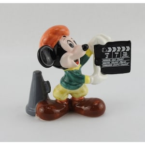 Peut inclure: Figurine en c&eacute;ramique de Mickey Mouse tenant un clap de cin&eacute;ma et un m&eacute;gaphone. Mickey porte une veste verte, un pantalon jaune et des chaussures marron. Le clap de cin&eacute;ma porte l'inscription "Sc&egrave;ne 2 Prise 1 Titre".