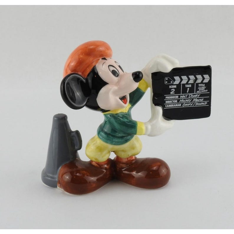 Peut inclure: Figurine en c&eacute;ramique de Mickey Mouse tenant un clap de cin&eacute;ma et un m&eacute;gaphone. Mickey porte une veste verte, un pantalon jaune et des chaussures marron. Le clap de cin&eacute;ma porte l'inscription "Sc&egrave;ne 2 Prise 1 Titre".