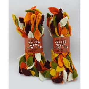 May include: Two bundles of handcrafted felted wool garland featuring leaf-shaped pieces in autumn colors including orange, yellow, dark red, olive green, and cream. Each bundle has a paper label reading Trader Joe's Handcrafted Felted Wool Garland 6ft Long.