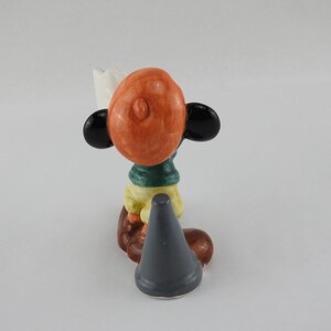Peut inclure: Figurine en c&eacute;ramique d'un personnage de dessin anim&eacute; avec une t&ecirc;te orange, des oreilles noires et une &eacute;charpe verte. La figurine porte une chemise jaune et un pantalon marron. Un objet gris en forme de c&ocirc;ne est devant la figurine.