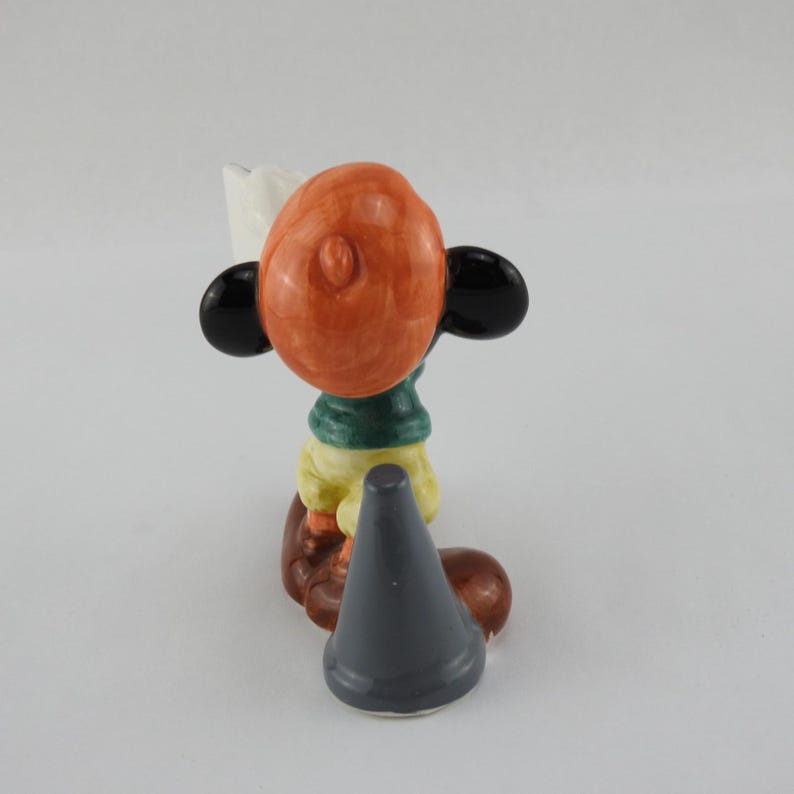 Peut inclure: Figurine en c&eacute;ramique d'un personnage de dessin anim&eacute; avec une t&ecirc;te orange, des oreilles noires et une &eacute;charpe verte. La figurine porte une chemise jaune et un pantalon marron. Un objet gris en forme de c&ocirc;ne est devant la figurine.
