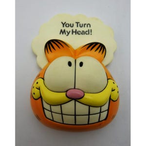 May include: A molded plastic Garfield character magnet featuring a bright orange face with large white eyes, a pink nose, and a wide yellow grin. Above the head is a cream-colored speech bubble shape with the black text, You Turn My Head!
