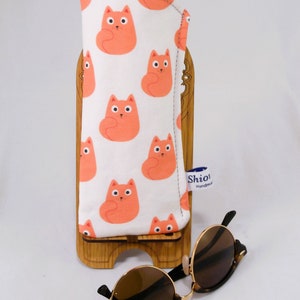 Puede incluir: Estuche para gafas de sol de tela con estampado de gatos blancos y naranjas con ribete negro. El estuche est&aacute; sobre un soporte de madera. Un par de gafas de sol de carey marr&oacute;n est&aacute;n sobre una superficie blanca.